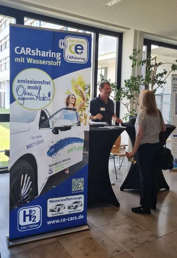 Ein Stand für Wasserstoff-Carsharing. Ein Mitarbeiter informiert eine interessierte Person über emissionsfreies Fahren ohne eigenes Auto. Im Hintergrund sind Pflanzen und ein hell erleuchteter Raum zu sehen. 