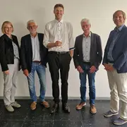 Eine Gruppe von fünf Personen steht aufrecht in einem Raum. In der Mitte hält ein großer Mann mit schwarzem Anzug ein Dokument in der Hand, während die anderen Personen in formeller Kleidung neben ihm stehen und in die Kamera schauen.