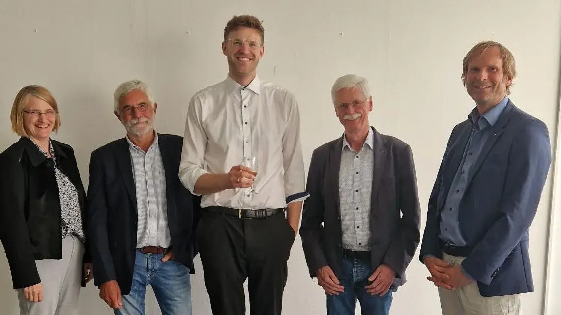 Eine Gruppe von fünf Personen steht aufrecht in einem Raum. In der Mitte hält ein großer Mann mit schwarzem Anzug ein Dokument in der Hand, während die anderen Personen in formeller Kleidung neben ihm stehen und in die Kamera schauen.