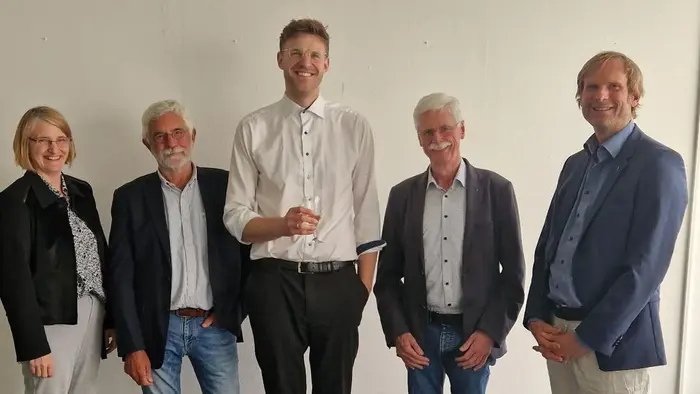 Eine Gruppe von fünf Personen steht aufrecht in einem Raum. In der Mitte hält ein großer Mann mit schwarzem Anzug ein Dokument in der Hand, während die anderen Personen in formeller Kleidung neben ihm stehen und in die Kamera schauen.