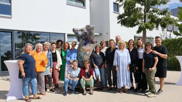 Eine Gruppe von 22 Menschen steht im Freien vor einem bunten Mosaikskulptur. Die Personen sind unterschiedlich gekleidet und lächeln. Im Hintergrund sieht man ein modernes Gebäude mit Fenstern und Bäumen.