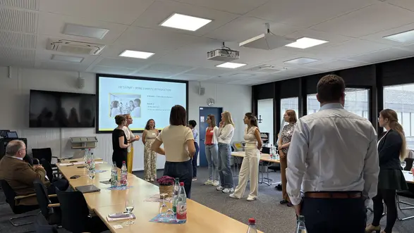 Eine Gruppe von Personen steht in einem Besprechungsraum und nimmt an einer Aktivität teil. Im Hintergrund ist ein Projektor mit einer Präsentation sichtbar. Auf dem Tisch sind Flaschen und Unterlagen verteilt. Die Stimmung ist interaktiv und kommunikativ.