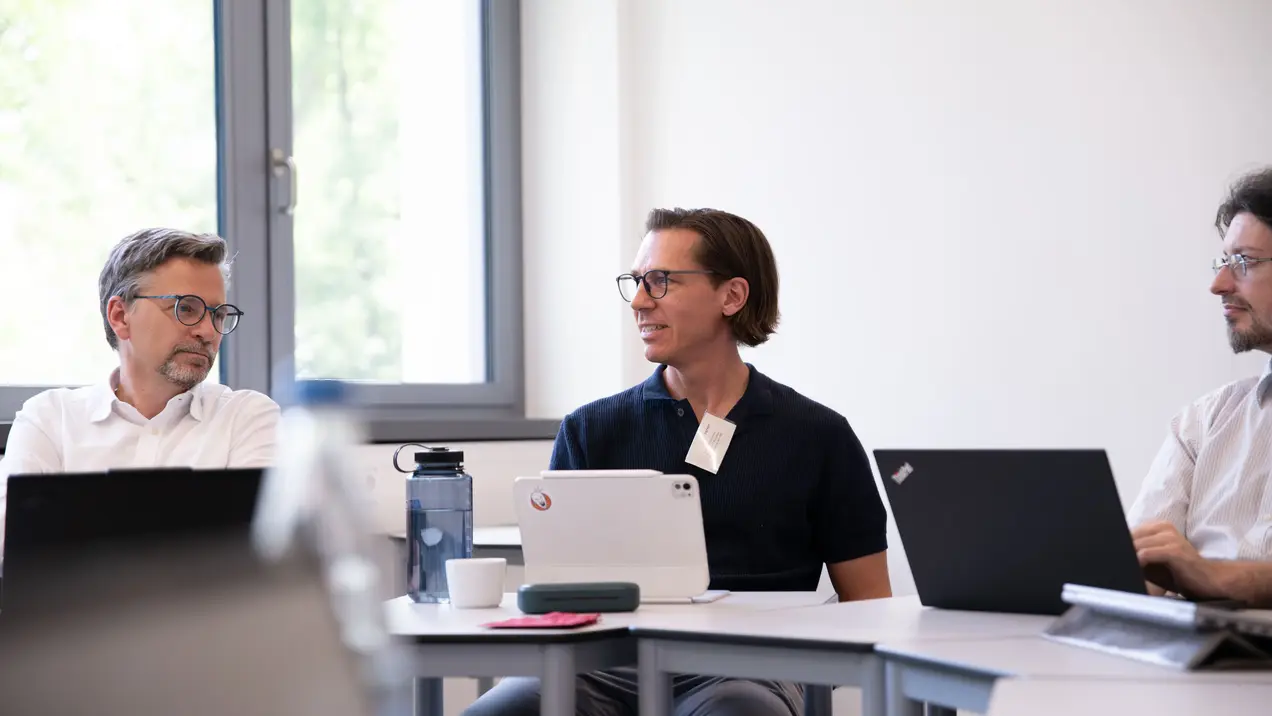 Drei Männer sitzen an einem Tisch in einem Besprechungsraum. Sie sind mit Laptops beschäftigt und führen ein offenes Gespräch. Der Raum hat viel Tageslicht, und eine freundliche, kreative Atmosphäre ist spürbar.