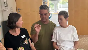 Eine Gruppe von drei Menschen sitzt in einem Raum. Zwei Personen, eine Frau in schwarzer Kleidung und ein Mann in grüner T-Shirt, diskutieren. Eine dritte Frau hört aufmerksam zu. Sie halten Blätter in der Hand und scheinen aktiv am Gespräch teilzunehmen.