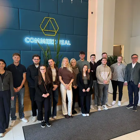 Eine Gruppe von zwölf Personen steht im Foyer eines Unternehmens mit der Aufschrift "COMMERZ REAL" im Hintergrund. Sie tragen verschiedene persönliche Stile und lächeln. Die Atmosphäre wirkt freundlich und einladend.