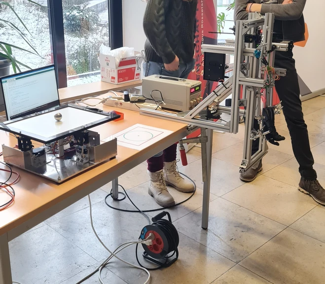 Ein Tisch mit elektronischen Geräten: ein Computer mit Bildschirm, eine Steuerungseinheit und ein mechanisches Gerät mit einem runden Tablett zur Bearbeitung. Zwei Personen stehen daneben und interagieren mit den Geräten. Im Hintergrund sind Fenster und Pflanzen sichtbar.