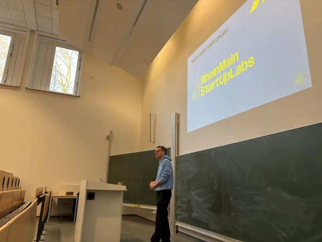Ein Referent steht an einem Rednerpult vor einer Präsentationsfolie mit dem Titel "RheinMain StartUpLabs". Im Hintergrund ist eine grüne Tafel sichtbar, und es gibt Fenster, durch die Tageslicht strömt. Der Raum wirkt nachdenklich und akademisch.