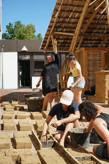 Four students are working on an outdoor construction project. They are making mud bricks using buckets and moulds. One person raises their hand in a cheerful gesture while the others concentrate on working with the material. A thatched roof can be seen in the background.