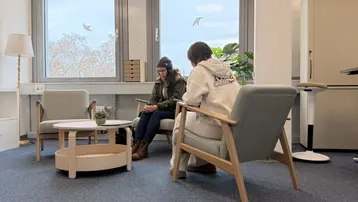 Zwei Personen sitzen in einem modernen Büro. Eine Person in einem grünen Mantel spricht mit einer Person in einem weißen Sweatshirt. Zwischen ihnen steht ein runder Tisch mit einer Pflanze. Das Fenster ist gut sichtbar und lässt Tageslicht herein.