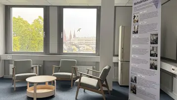 Ein heller Raum mit grauem Teppichboden. In der Mitte stehen drei Stühle und ein runder Tisch. An einer Wand befindet sich ein Informationsbanner zum Thema "Mendenlabor" mit Texten und Bildern. Durch die Fenster ist eine grüne Baumlandschaft sichtbar.