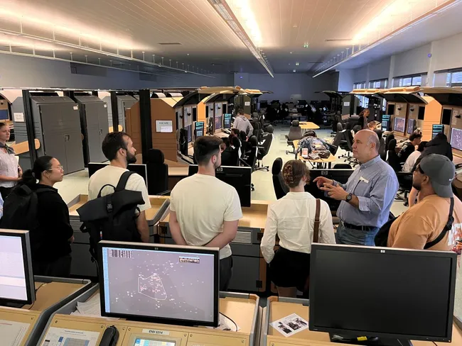 Eine Gruppe von Personen lauscht einem Vortrag eines Dozenten in einem Schulungsraum mit mehreren Computern und Arbeitsplätzen. Im Hintergrund sind weitere Teilnehmer und technische Geräte zu sehen. Die Atmosphäre ist informativ und bildungsorientiert.