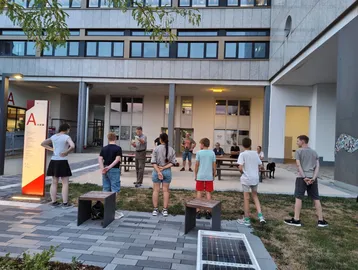 Eine Gruppe von Kindern steht im Freien und hört aufmerksam einem älteren Mann zu, der ihnen etwas erklärt. Im Hintergrund sind Bänke und ein modernes Gebäude sichtbar. Die Szene wirkt informativ und gemeinschaftlich.