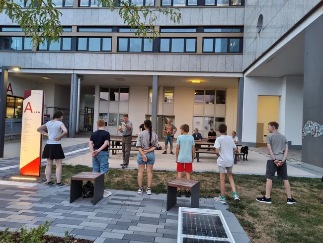 Eine Gruppe von Kindern steht im Freien und hört aufmerksam einem älteren Mann zu, der ihnen etwas erklärt. Im Hintergrund sind Bänke und ein modernes Gebäude sichtbar. Die Szene wirkt informativ und gemeinschaftlich.