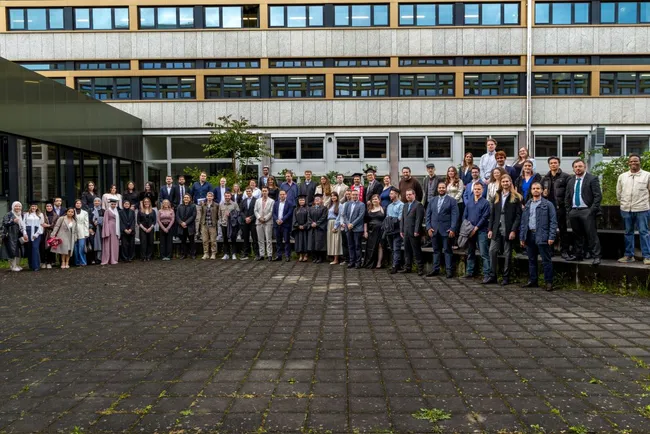 Gruppenfoto von über 50 Personen, die im Freien stehen. Die Personen tragen formelle Kleidung und lächeln in die Kamera. Im Hintergrund ist ein modernes, mehrstöckiges Gebäude mit großen Fenstern zu sehen, umgeben von einem gepflasterten Platz.