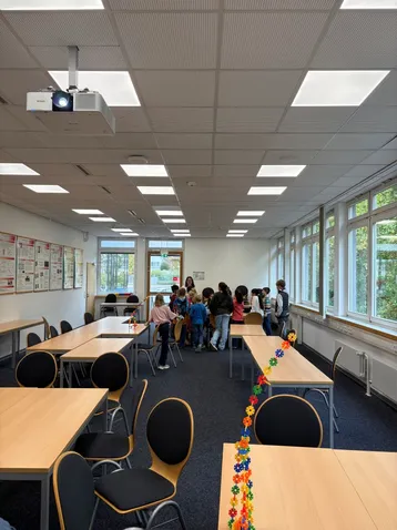 In einem hellen Raum stehen mehrere Kinder um einen Tisch. Tische sind leer und der Raum hat große Fenster, die Licht hereinlassen. An der Decke hängt ein Projektor. Die Atmosphäre ist lebhaft und lernfördernd.