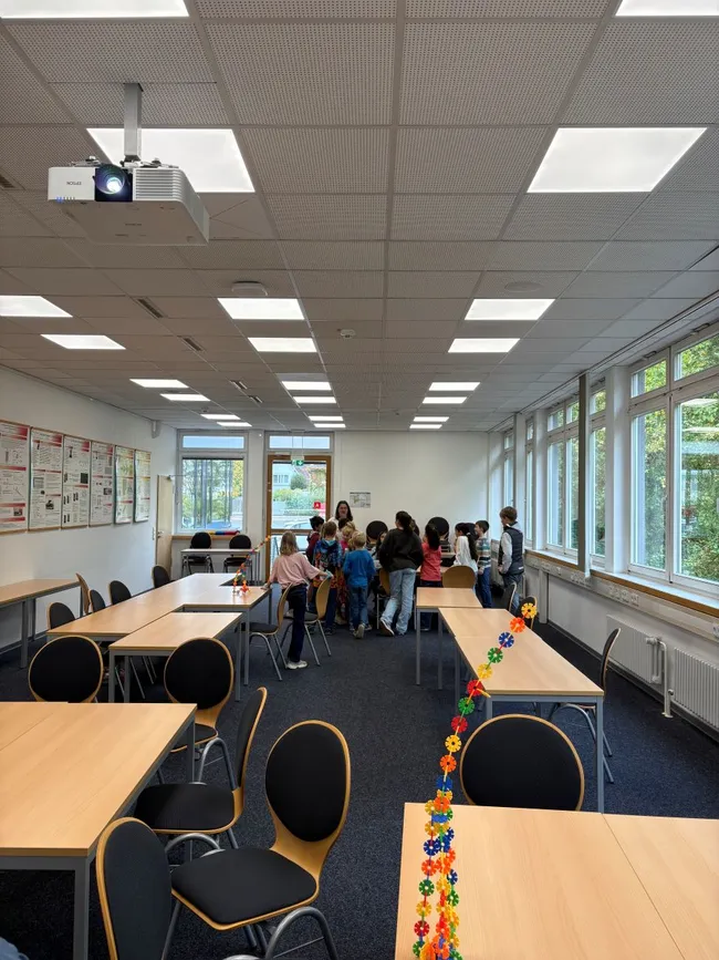 In einem hellen Raum stehen mehrere Kinder um einen Tisch. Tische sind leer und der Raum hat große Fenster, die Licht hereinlassen. An der Decke hängt ein Projektor. Die Atmosphäre ist lebhaft und lernfördernd.