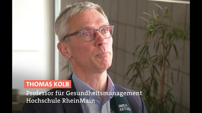 Ein Mann mit Brille und grauen Haaren spricht in die Kamera. Er ist Thomas Kolb, Professor für Gesundheitsmanagement an der Hochschule RheinMain. Im Hintergrund sind unscharfe Elemente eines Raumes sichtbar.