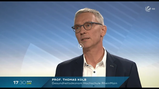 Ein Mann mit Brille und kurzem, grauem Haar spricht in ein Mikrofon. Er trägt einen dunklen Anzug und sein Gesichtsausdruck ist nachdenklich. Im Hintergrund ist ein sanfter Farbverlauf zu sehen. Unter ihm steht sein Name und Titel: „Prof. Thomas Kolb, Gesundheitsökonom Hochschule RheinMain“.