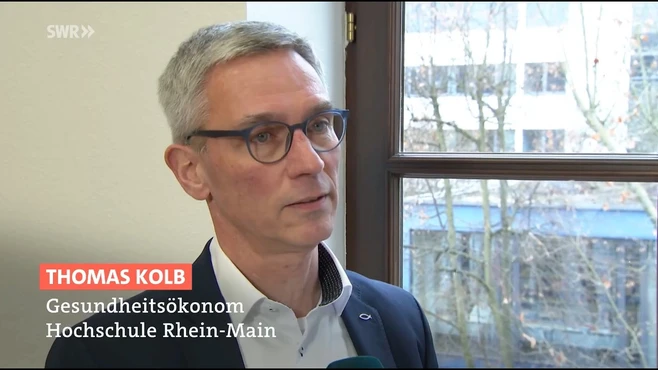 Ein Mann spricht in einem Interview. Er trägt eine Brille und hat kurze Haare. Zu sehen sind Teile eines Fensters im Hintergrund, durch das Tageslicht strömt. Neben ihm steht der Text: "Thomas Kolb, Gesundheitsökonom, Hochschule Rhein-Main."