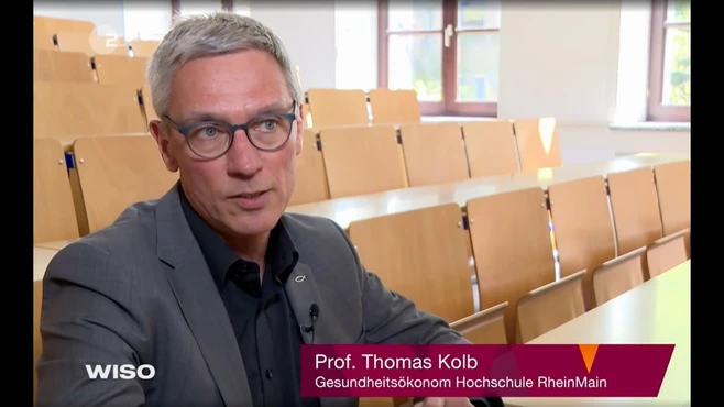 Ein Mann mit Brille, Professor Thomas Kolb, sitzt in einem leeren Klassenraum. Er trägt ein schwarzes Hemd und spricht über Gesundheitsökonomie an der Hochschule RheinMain. Auf dem Bildschirm wird sein Name und Titel angezeigt.