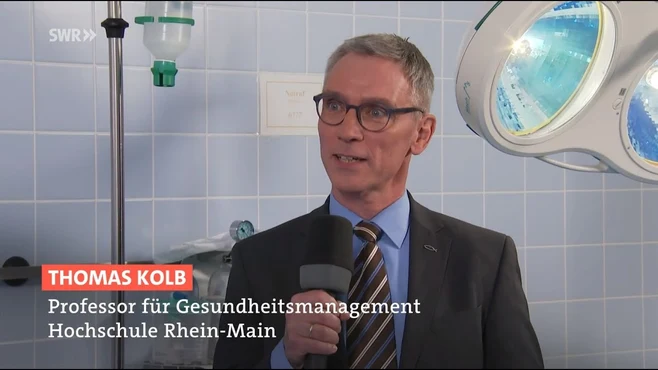 Ein Mann mit Brille und Anzug spricht in einem medizinischen Umfeld. Er stellt sich als Thomas Kolb vor, Professor für Gesundheitsmanagement an der Hochschule Rhein-Main. Im Hintergrund sind Operationslichter zu sehen.