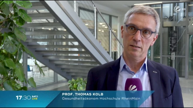 Ein Mann mit Brille und grauen Haaren steht in einem modernen Gebäude. Er trägt ein dunkles Jackett und spricht in ein Mikrofon. Unter ihm steht „Prof. Thomas Kolb, Gesundheitsökonom, Hochschule RheinMain“ und die Uhrzeit „17:30 SAT1 LIVE“ im unteren Bildbereich.