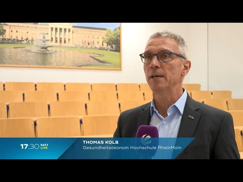 Ein Mann namens Thomas Kolb, Gesundheitsökonom von der Hochschule RheinMain, steht in einem leerem Seminarraum. Er spricht in die Kamera, während im Hintergrund ein Bild eines Brunnens und eines Gebäudes sichtbar ist.