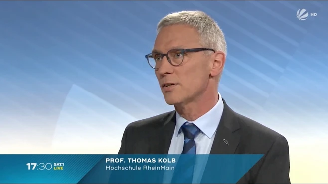 Ein Professor spricht in einem TV-Studio. Er trägt eine Brille, ein dunkles Sakko und eine helle Krawatte. Im unteren Bereich steht sein Name und seine Zugehörigkeit zur Hochschule RheinMain. Der Hintergrund ist hell und leicht strukturiert.